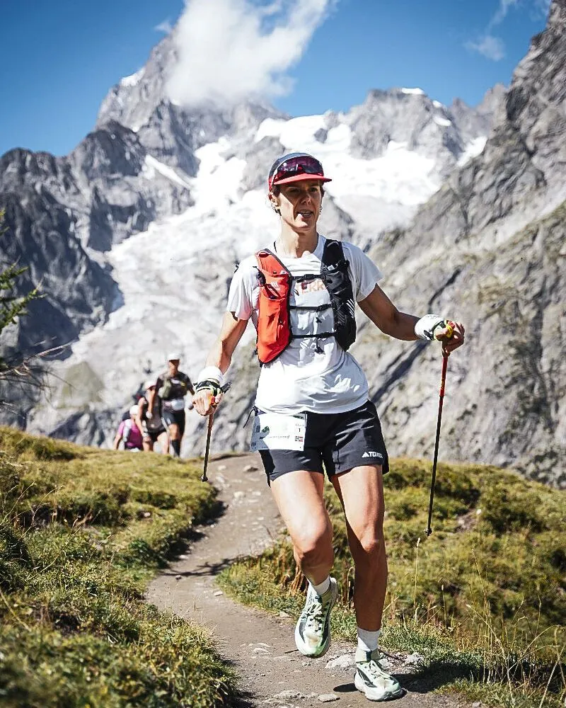 Anne Mjelva på vei oppover en sti i Alpene med staver, med snødekte tinder bak seg.