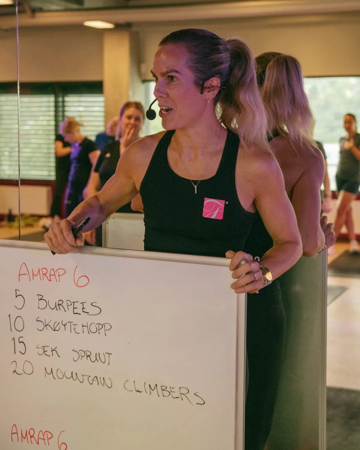 Anne Mjelva leading a class with a headset microphone, holding up a board with the session: AMRAP 6, 5 burpees, 10 skater jumps, 15 sec sprint, 20 mountain climbers.